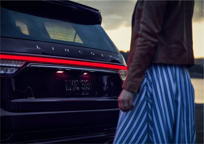 A person is shown near the rear of a 2023 Lincoln Aviator® SUV as the Lincoln Embrace illuminates the rear lights | Pugmire Lincoln of Marietta in Marietta GA